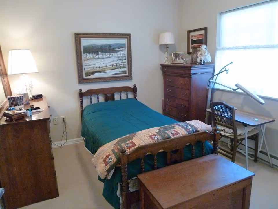 Small furnished bedroom with a wooden single bed covered in a green comforter, dressers, a desk by the window, and framed artwork on the wall.