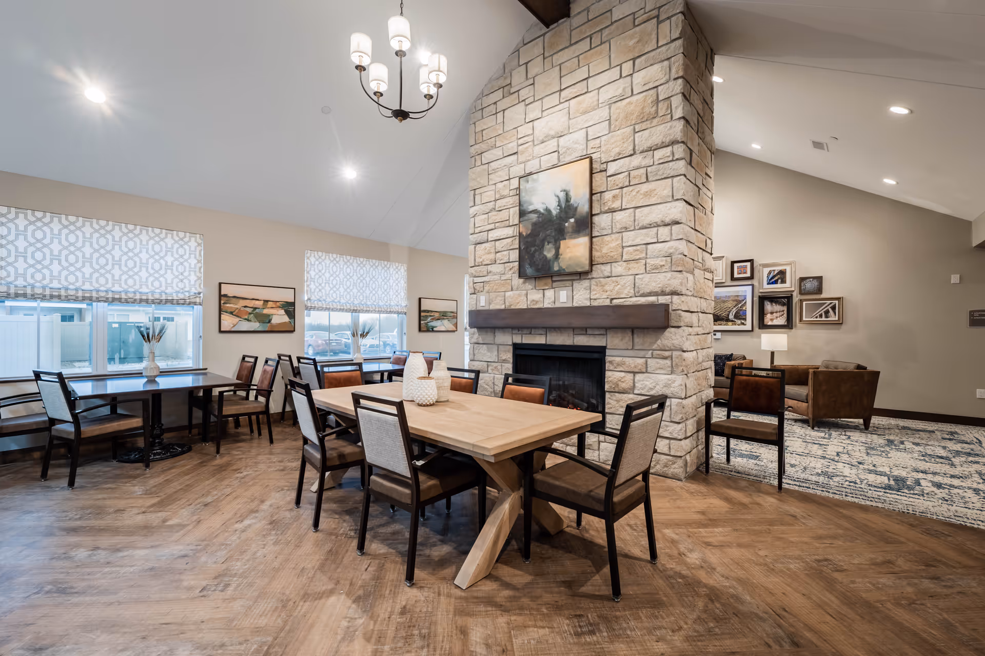 A senior living facility common area featuring a stone fireplace in the center with a wooden mantel and an abstract painting above it. Surrounding the fireplace are several tables and chairs arranged for dining or socializing. The room has large windows with patterned blinds, wooden flooring, and a chandelier hanging from the ceiling. In the background, there is a seating area with armchairs and framed pictures on the wall.