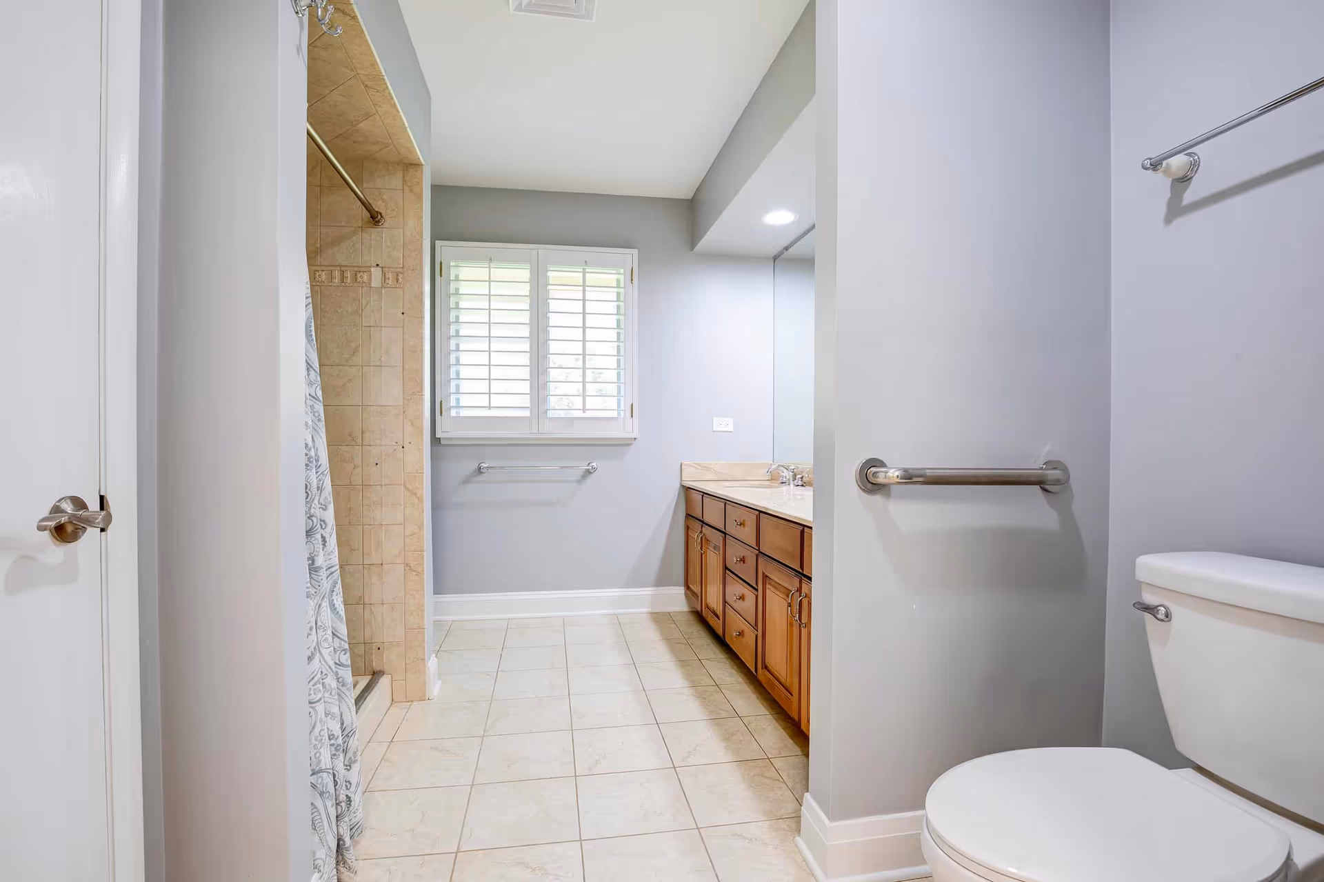 Bright bathroom with beige tiled floor and shower area with a curtain on the left. There is a large wooden vanity with multiple drawers and a countertop with a sink on the right side. A white toilet is visible in the foreground on the right. The walls are painted light gray, and there is a window with white plantation shutters above a towel rack on the far wall.