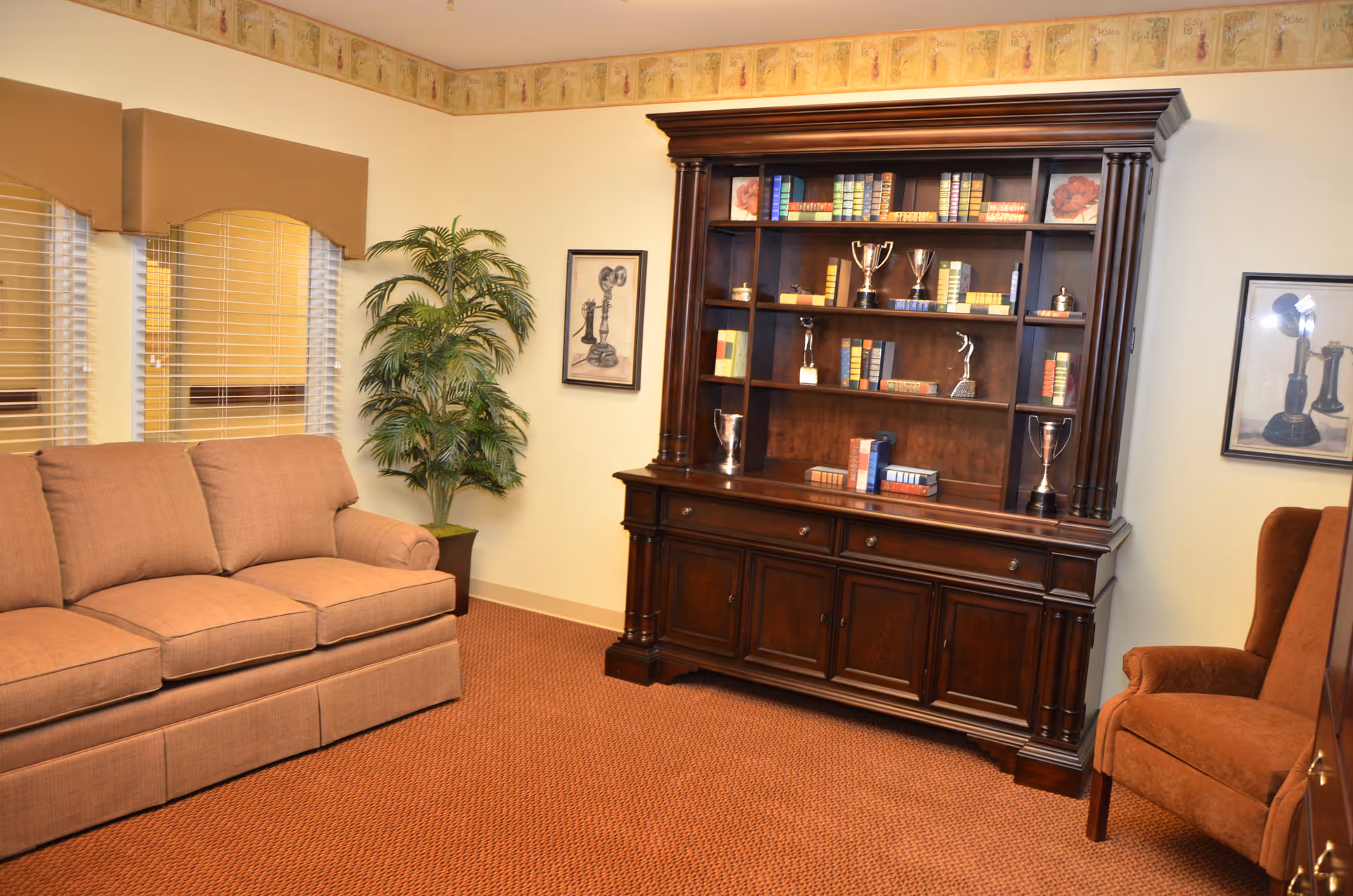 Cozy sitting room with a beige sofa, large wooden bookcase displaying books and trophies, a potted plant, and an armchair.