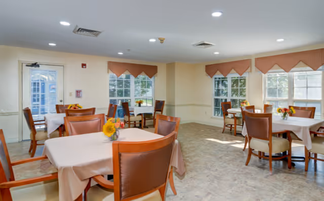 Bright dining room with several tables covered in tablecloths and chairs, each table decorated with a small flower arrangement and large windows letting in natural light.