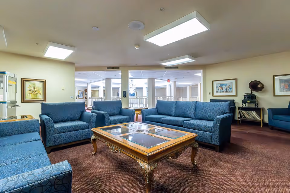 A spacious living room area with multiple blue upholstered sofas arranged around a wooden coffee table with glass panels. The room has beige walls adorned with framed artwork and a vintage gramophone on a side table. The ceiling has several rectangular fluorescent lights, and the floor is carpeted in a dark reddish-brown color. In the background, there is an open area with white pillars and railings.