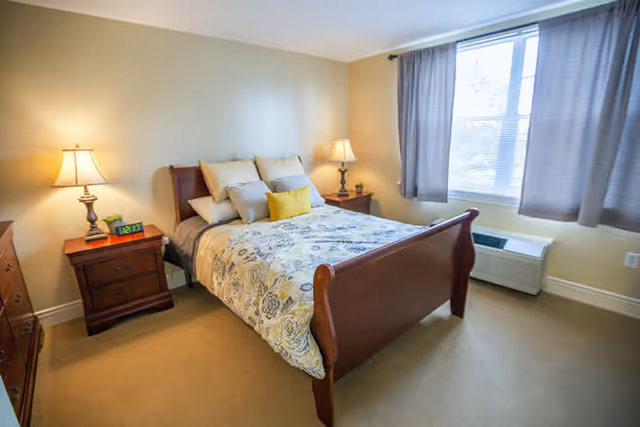 A cozy bedroom with a wooden bed frame, a floral patterned bedspread, multiple pillows, two bedside tables each with a lamp, a digital alarm clock on the left table, a window with gray curtains, and a heating/cooling unit below the window.