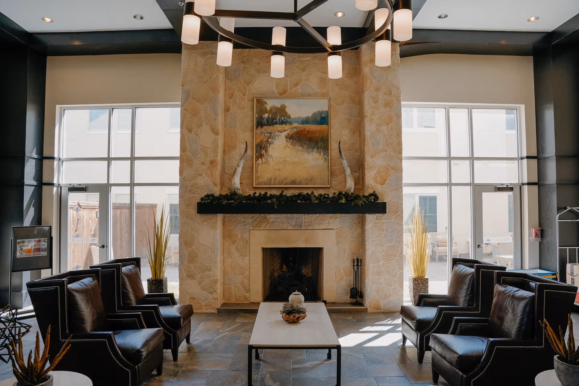 A cozy common area with a stone fireplace as the focal point, flanked by large windows letting in natural light. The fireplace mantel is decorated with greenery and two white horn-like sculptures, with a landscape painting hanging above. Four dark leather armchairs are arranged around a rectangular coffee table with a decorative centerpiece. The room has a modern chandelier hanging from the ceiling and plants placed near the windows.
