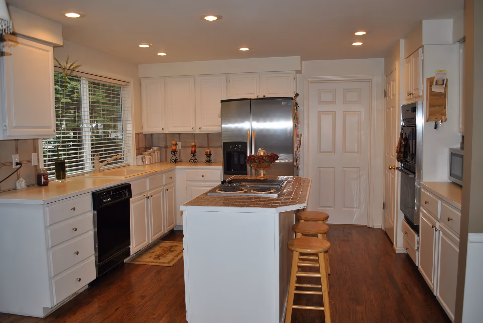 A bright kitchen with white cabinetry, a large window with blinds, a stainless steel refrigerator, a built-in oven, a microwave, and a kitchen island with a tiled countertop and three wooden stools. The floor is wooden, and there are recessed ceiling lights illuminating the space.