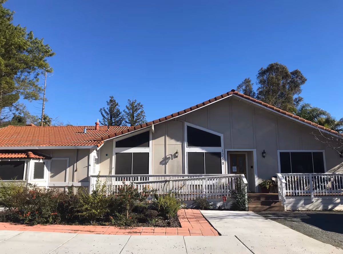 Single-story light-colored house with a red tile roof, front porch and ramp with white railings, and landscaping under a clear blue sky.