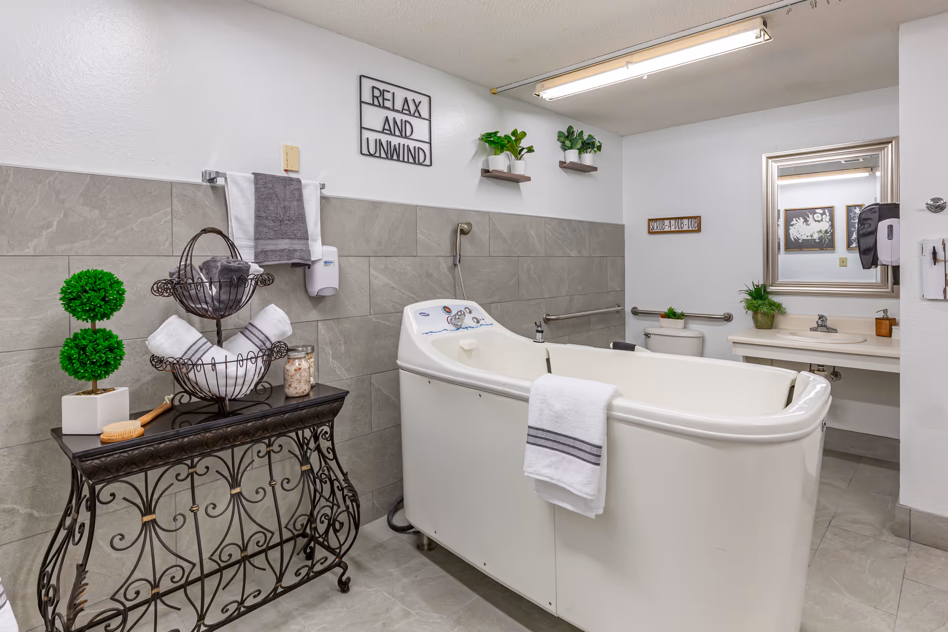 Accessible bathroom featuring a walk-in bathtub, sink and toilet, decorative table with towels and plants, and a 'Relax and Unwind' wall sign.