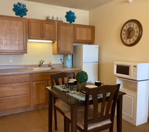 A small kitchen area with wooden cabinets, a white refrigerator, a microwave on a white stand, and a wooden dining table set with four chairs. The table has place settings and a small green plant centerpiece. A round wall clock is mounted on the beige wall above the microwave.