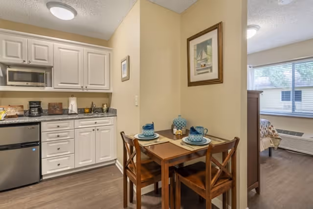 Kitchenette and dining nook featuring white cabinets, a stainless microwave and mini-fridge, and a two-person wooden table set with blue dishware beside an opening to a bedroom.