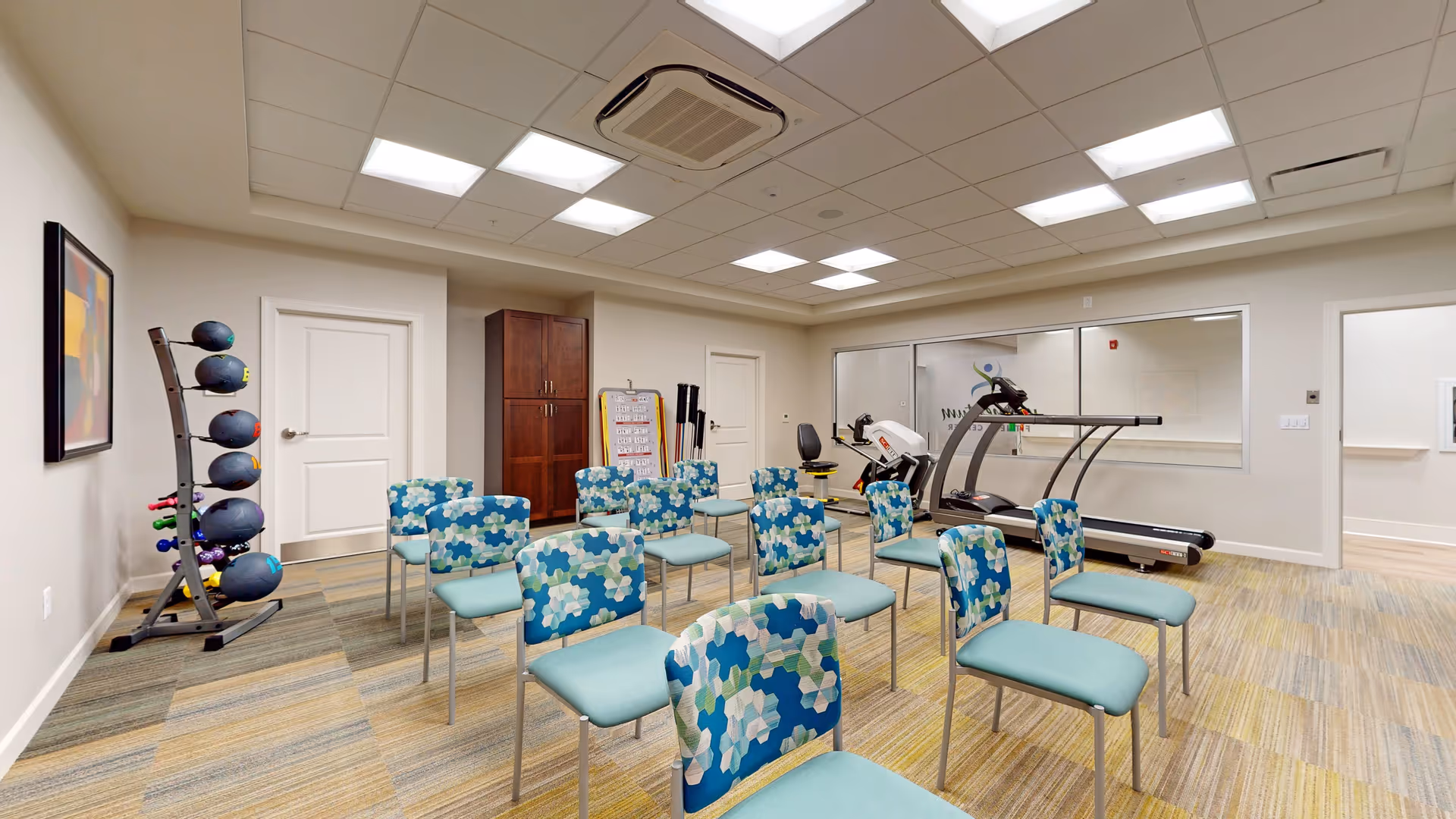 A well-lit exercise room with multiple chairs arranged in rows, a treadmill, exercise equipment including medicine balls and dumbbells on racks, a wooden cabinet, and a whiteboard. The room has a patterned carpet and a drop ceiling with fluorescent lights.
