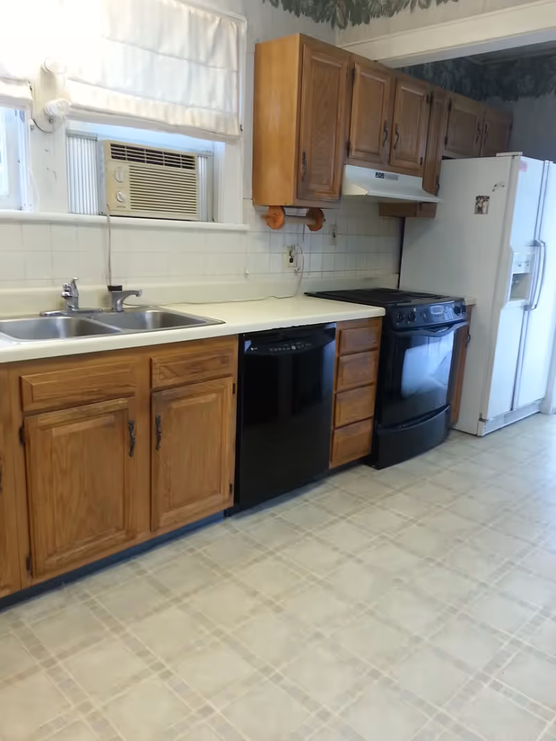 A kitchen with wooden cabinets, a double sink, a black dishwasher, a black stove with an oven, and a white refrigerator. There is a window above the sink with a white curtain and an air conditioning unit installed. The floor has a light-colored tile pattern.