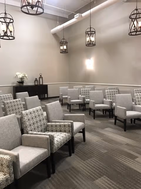 A spacious room with multiple rows of upholstered chairs arranged facing forward. The chairs have a mix of solid light gray and patterned upholstery. The room features beige walls, a carpeted floor with a striped pattern, and several hanging lantern-style light fixtures from the ceiling. A small black cabinet with decorative items and a white flower arrangement is visible against the back wall.