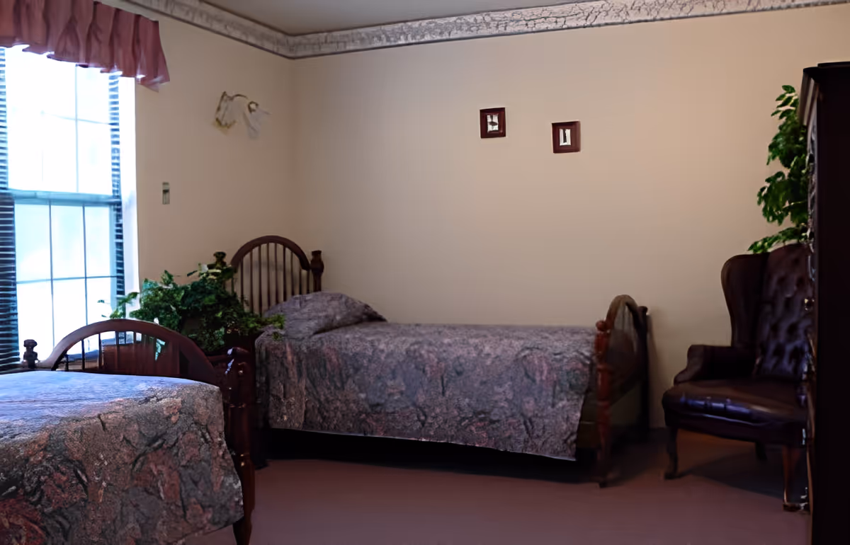 Two twin beds in a softly lit bedroom with a window, potted plants, and a leather armchair.