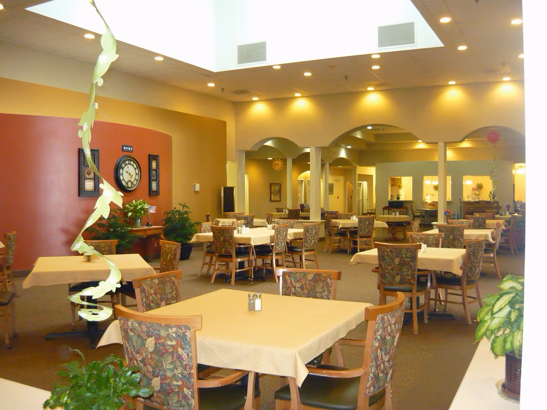 A spacious dining room with multiple tables covered in beige tablecloths and surrounded by floral upholstered chairs. The room features warm lighting, a large clock on a red accent wall, potted plants, and decorative hanging streamers. The ceiling has recessed lighting and a skylight, creating a bright and welcoming atmosphere.