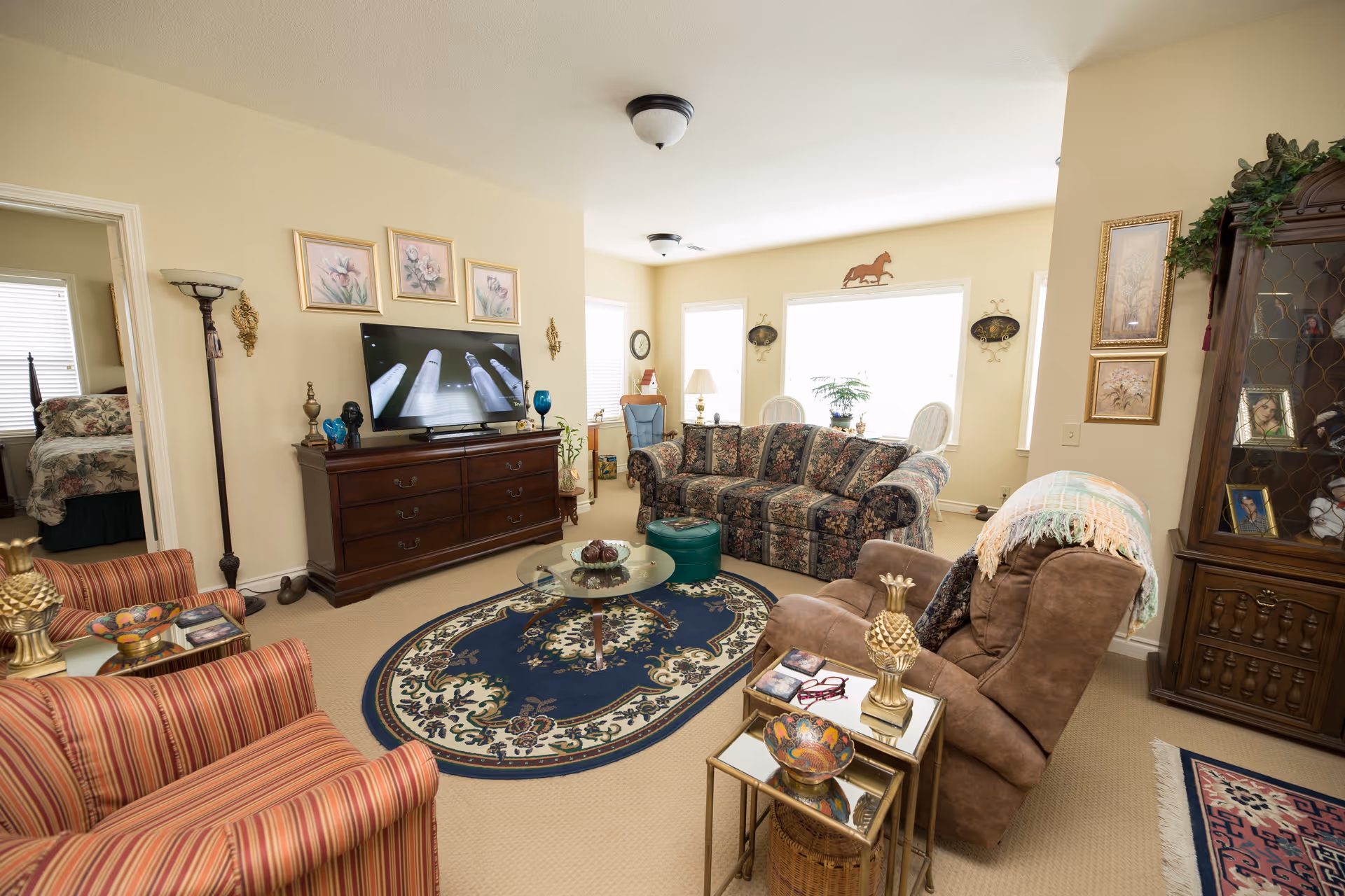 A cozy living room with a floral patterned sofa, a brown recliner with a throw blanket, and two striped armchairs. A wooden dresser with a flat-screen TV on top is against one wall, with three framed floral paintings above it. A glass coffee table sits on an oval patterned rug in the center of the room. The room is well-lit with natural light coming from large windows, and there is a wooden display cabinet with framed photos and decorative items on the right side.