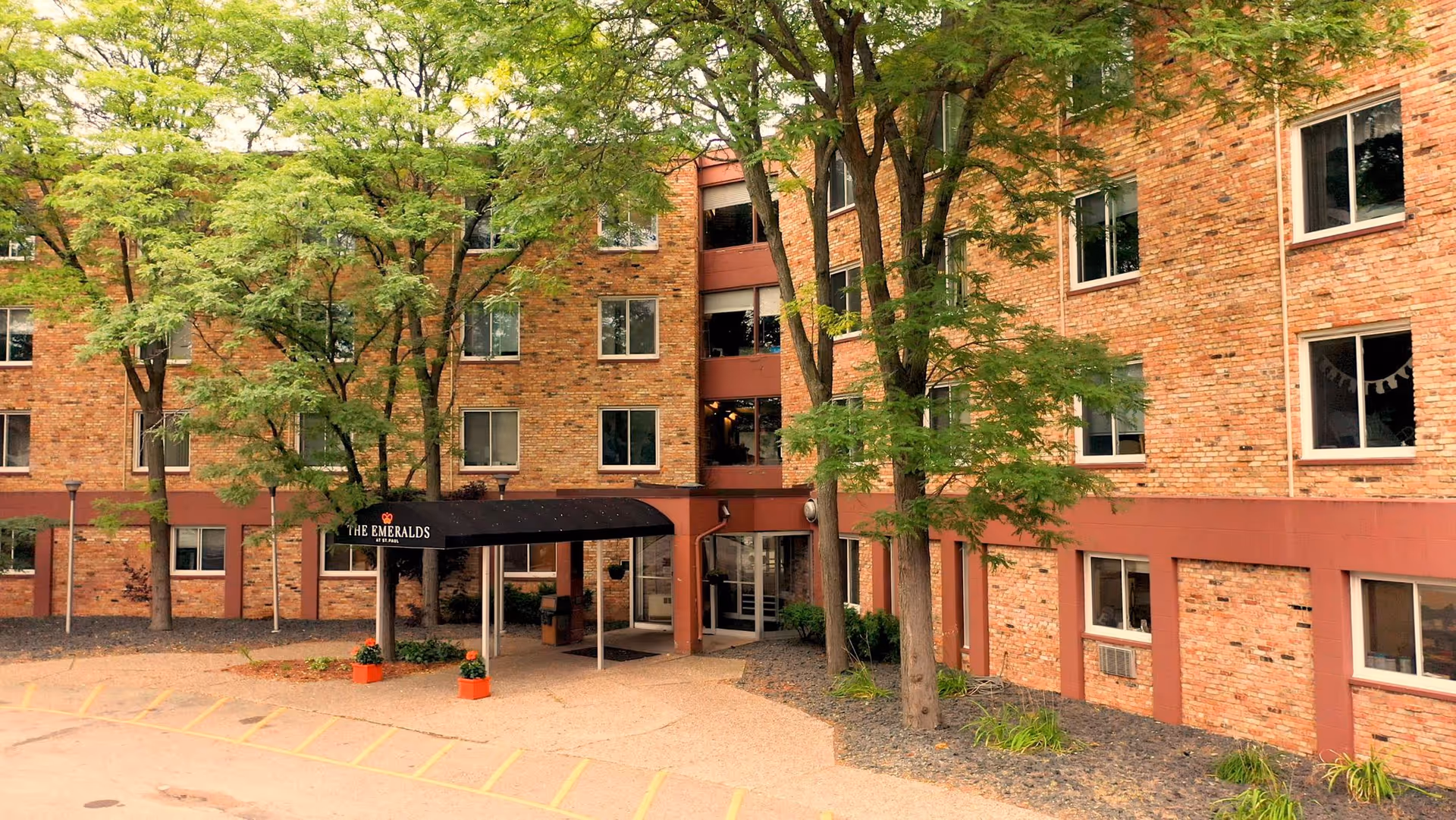 Exterior view of a multi-story brick building with several windows, trees in front, and a covered entrance with a black awning that reads 'The Emeralds'. There are small plants and a paved area with parking lines visible.