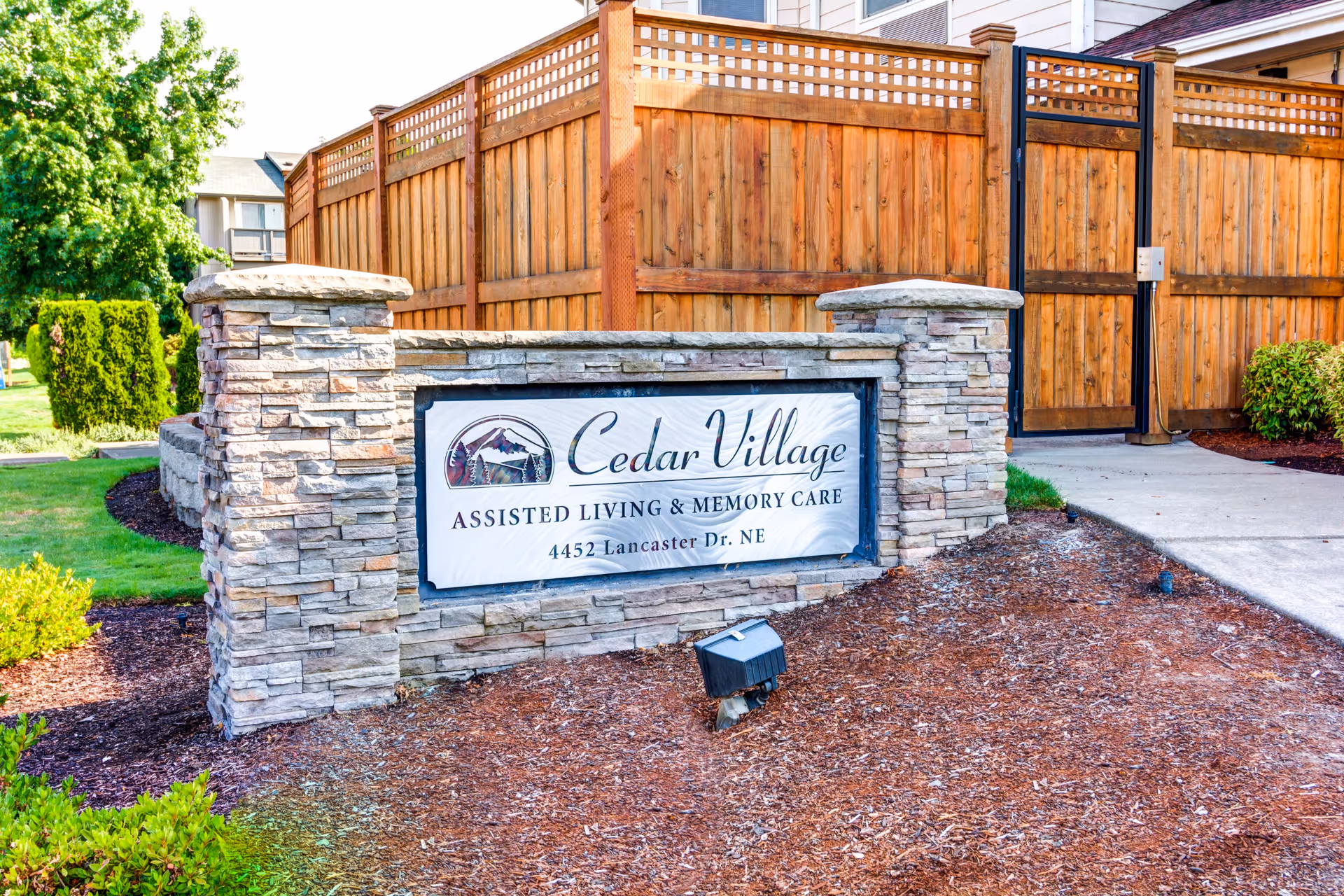 Stone sign for Cedar Village Assisted Living & Memory Care located at 4452 Lancaster Dr. NE, set in a landscaped area with mulch, bushes, and a wooden fence in the background.