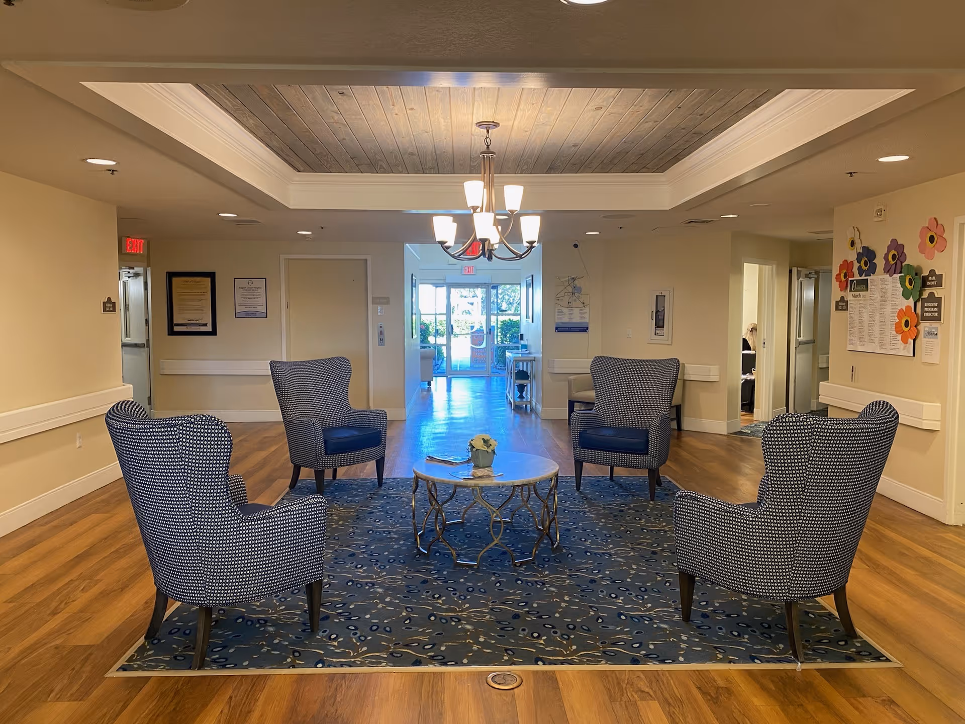 A seating area in a senior living facility with four patterned armchairs arranged around a round coffee table on a blue patterned rug. The room has wooden flooring, beige walls, and a wooden ceiling inset with a chandelier. There are doors and bulletin boards on the walls, and a hallway leading to a glass exit door with natural light coming through.