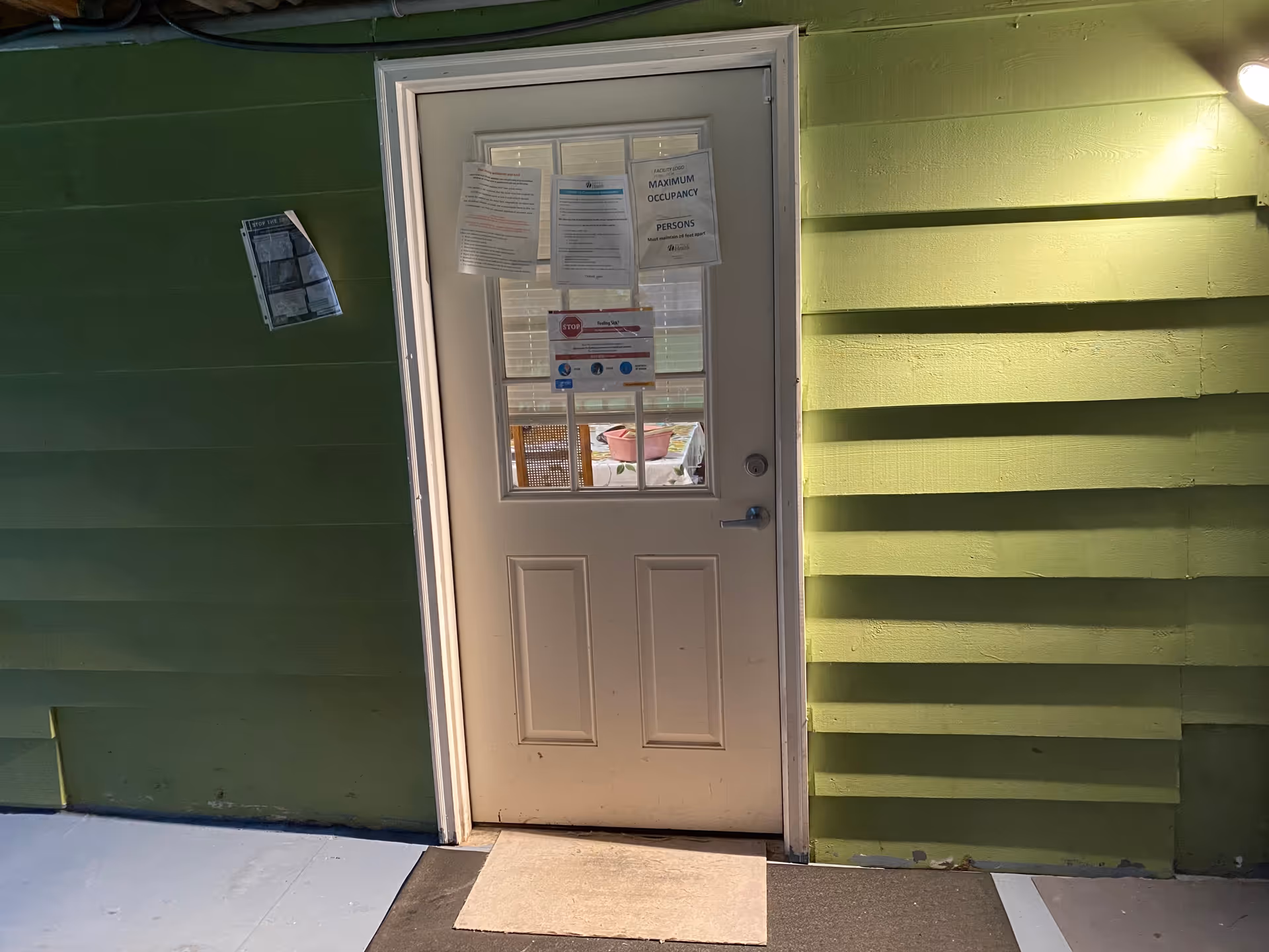 A white exterior door with a small window and posted notices set in bright green siding under an outdoor light.