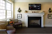 A cozy living room area with two patterned armchairs near a large window on the left. A white fireplace with a black interior is centered on the wall, above which a flat-screen TV is mounted. The mantel is decorated with small pumpkins and other fall-themed items. Two wall sconces with candles flank the fireplace, and framed pictures hang on either side.