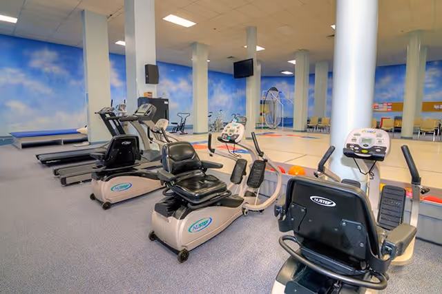 Interior view of a fitness room in a senior living facility with exercise equipment including recumbent bikes and treadmills. The room has a blue sky mural on the walls, several white columns, and a seating area with chairs in the background.