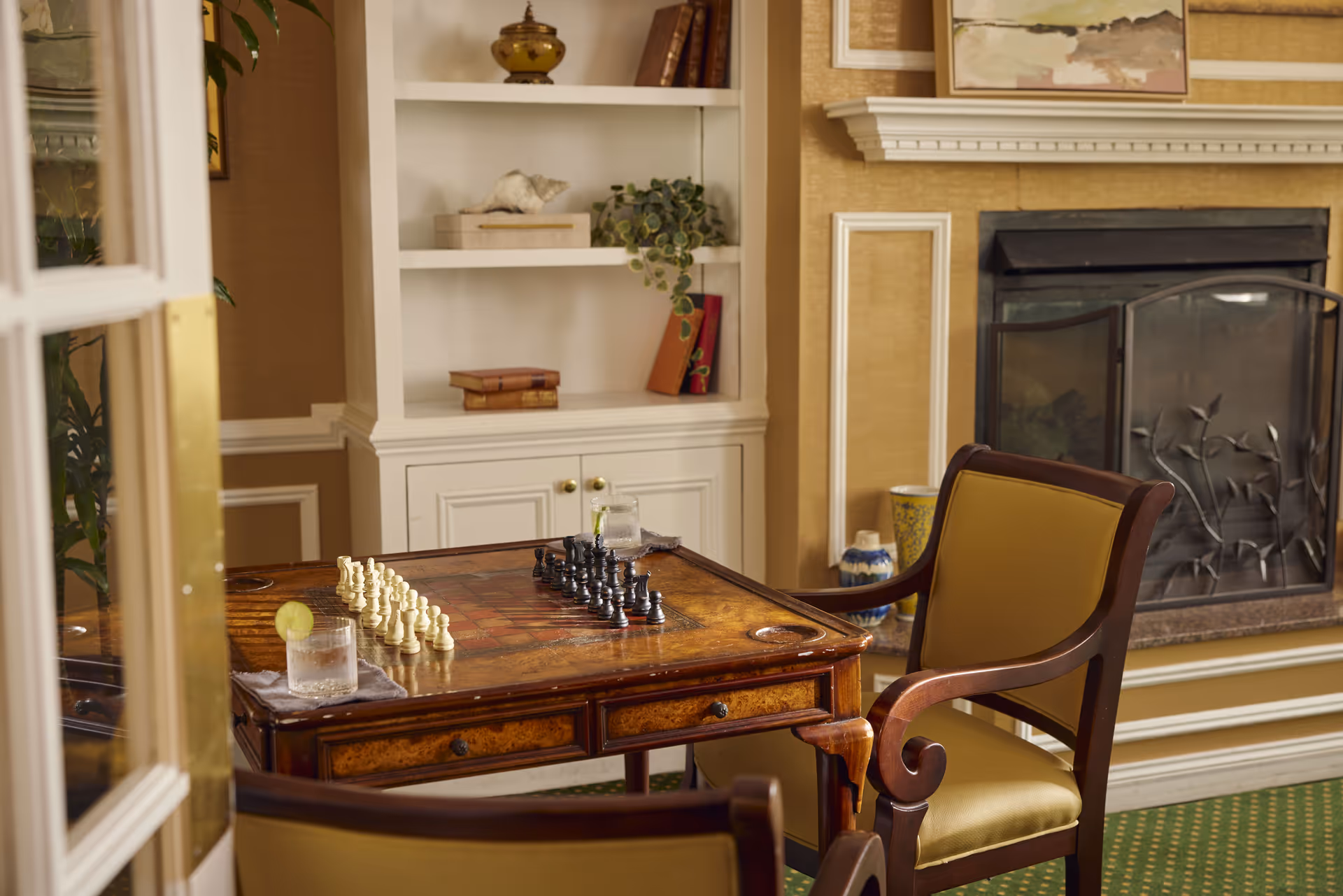 A cozy room with a wooden chess table set up for a game, two cushioned wooden chairs, a fireplace with a decorative screen, and a white built-in bookshelf with books and plants.