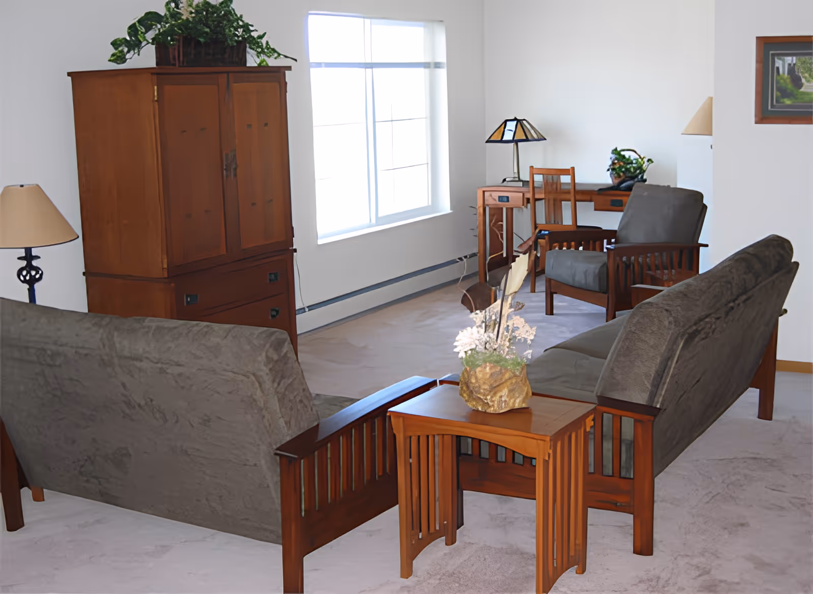 A cozy living room with two gray cushioned wooden sofas facing each other, a wooden side table with a floral arrangement, a wooden cabinet with a plant on top, a desk with a chair and a lamp, and a large window letting in natural light.