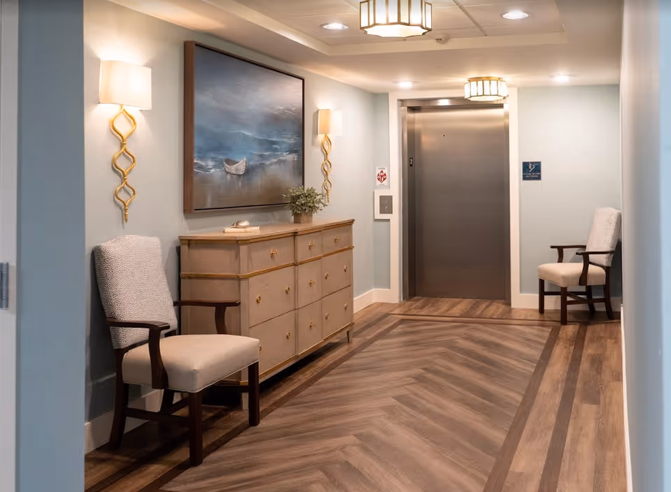Interior hallway of a senior living facility with a wooden floor, two upholstered chairs on either side, a wooden dresser with decorative items, wall sconces, a large painting of a boat on water, and a closed elevator door at the end.