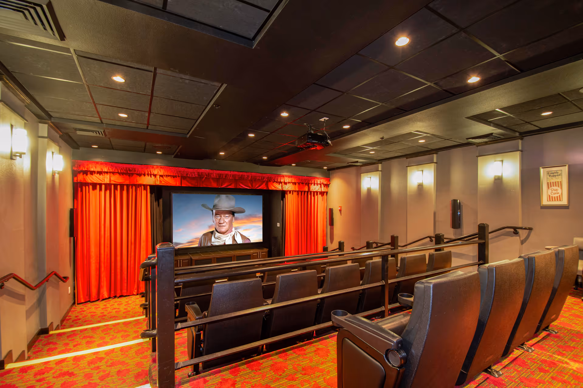 Interior view of a small movie theater room with red carpet and red curtains around the screen. The screen displays an image of a man wearing a cowboy hat and scarf. The room has several rows of black leather theater seats with cup holders and wall-mounted lights.