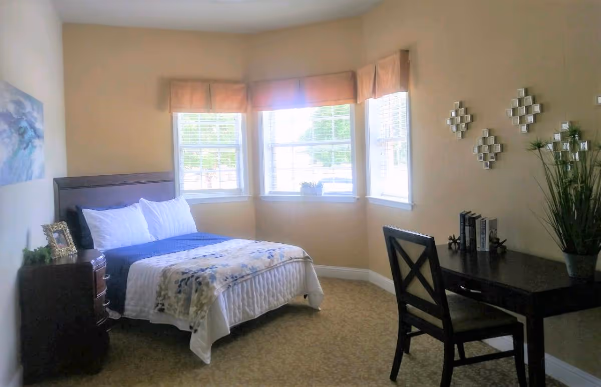 Well-lit bedroom with a made bed, nightstand, desk and chair beside a bay window.
