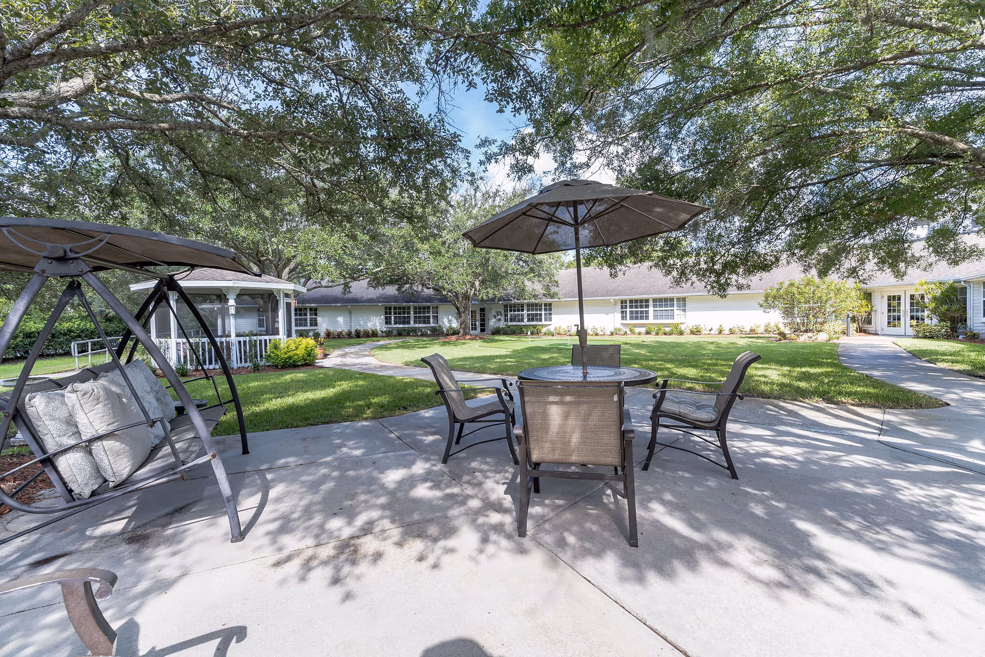 Outdoor patio area at The Landmark featuring a round table with an umbrella and four chairs, a cushioned swing, surrounded by green grass, trees providing shade, and a white building in the background.