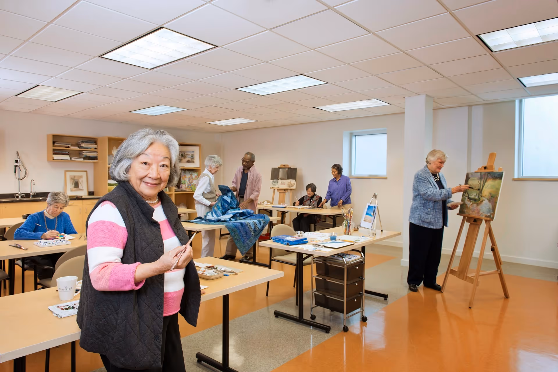 A group of elderly people engaged in various art activities in a bright, spacious room. One woman in the foreground smiles at the camera holding a paintbrush. Others are painting on canvases, working on crafts, or interacting with each other. The room has tables, chairs, shelves with art supplies, and large windows letting in natural light.