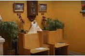 Interior view of a small chapel or prayer room with wooden pews, a wall clock, framed pictures, potted plants, and a religious statue on a shelf against a yellow wall.