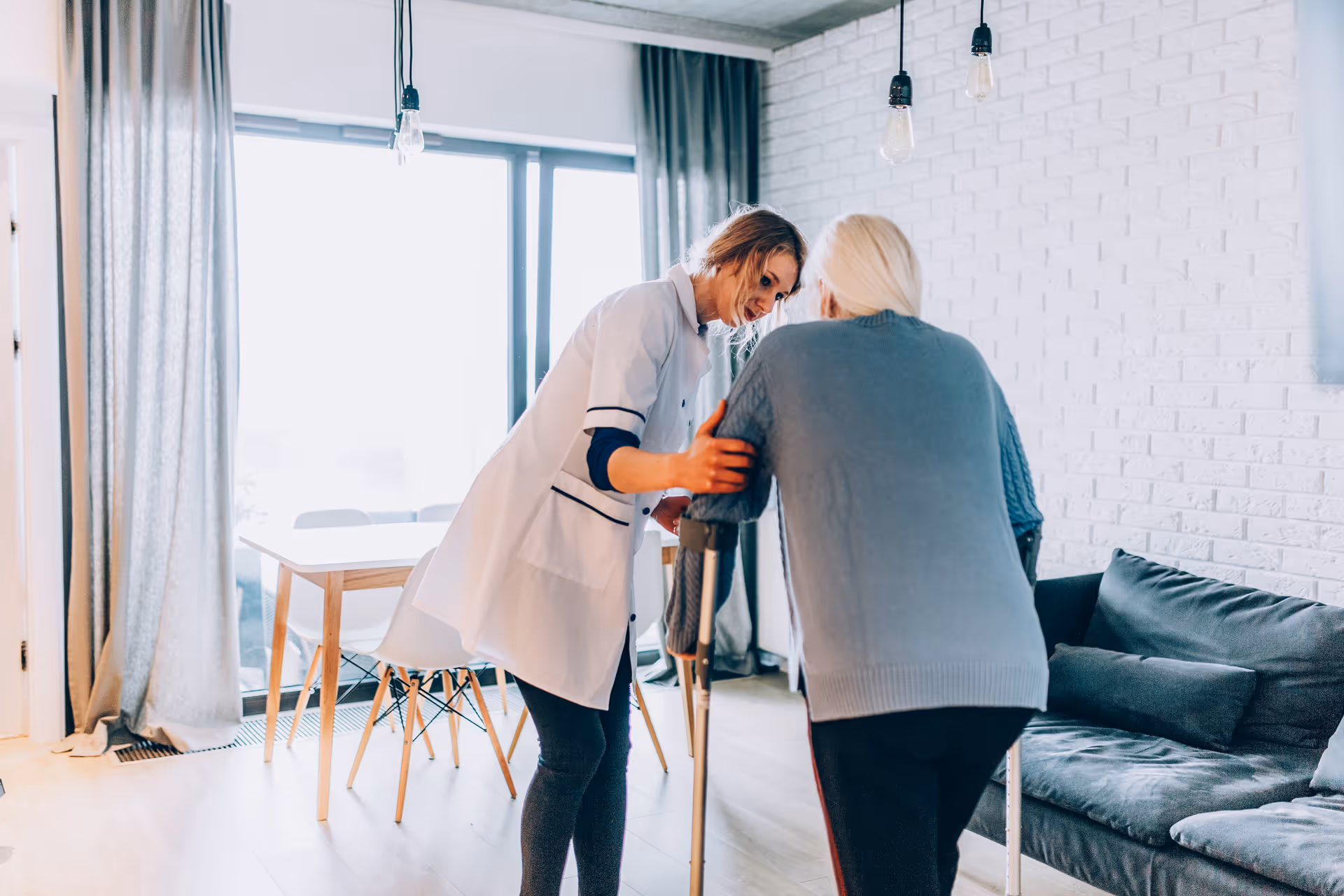 A caregiver assists an elderly woman using crutches in a bright living room with a gray couch, white brick wall, and a dining table near large windows with curtains.
