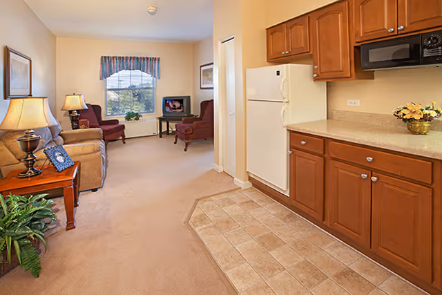 Interior view of a senior living facility apartment showing a small kitchen area with wooden cabinets, a white refrigerator, and a microwave. Adjacent to the kitchen is a living room with beige carpet, a sofa, two armchairs, a side table with a lamp, a TV on a stand, and a window with a valance letting in natural light.