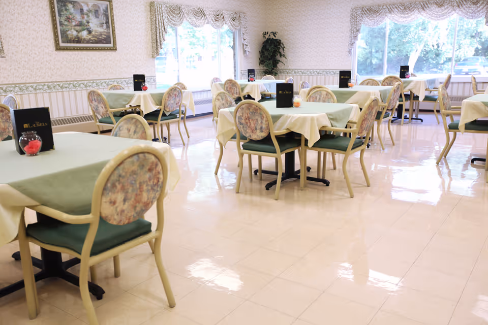 A bright dining room with multiple tables covered with green and white tablecloths. Each table is surrounded by chairs with floral patterned backs and green seats. Menus labeled 'Laurels' and small decorative items are placed on the tables. Large windows with floral valances let in natural light, and a framed painting hangs on the wallpapered wall.