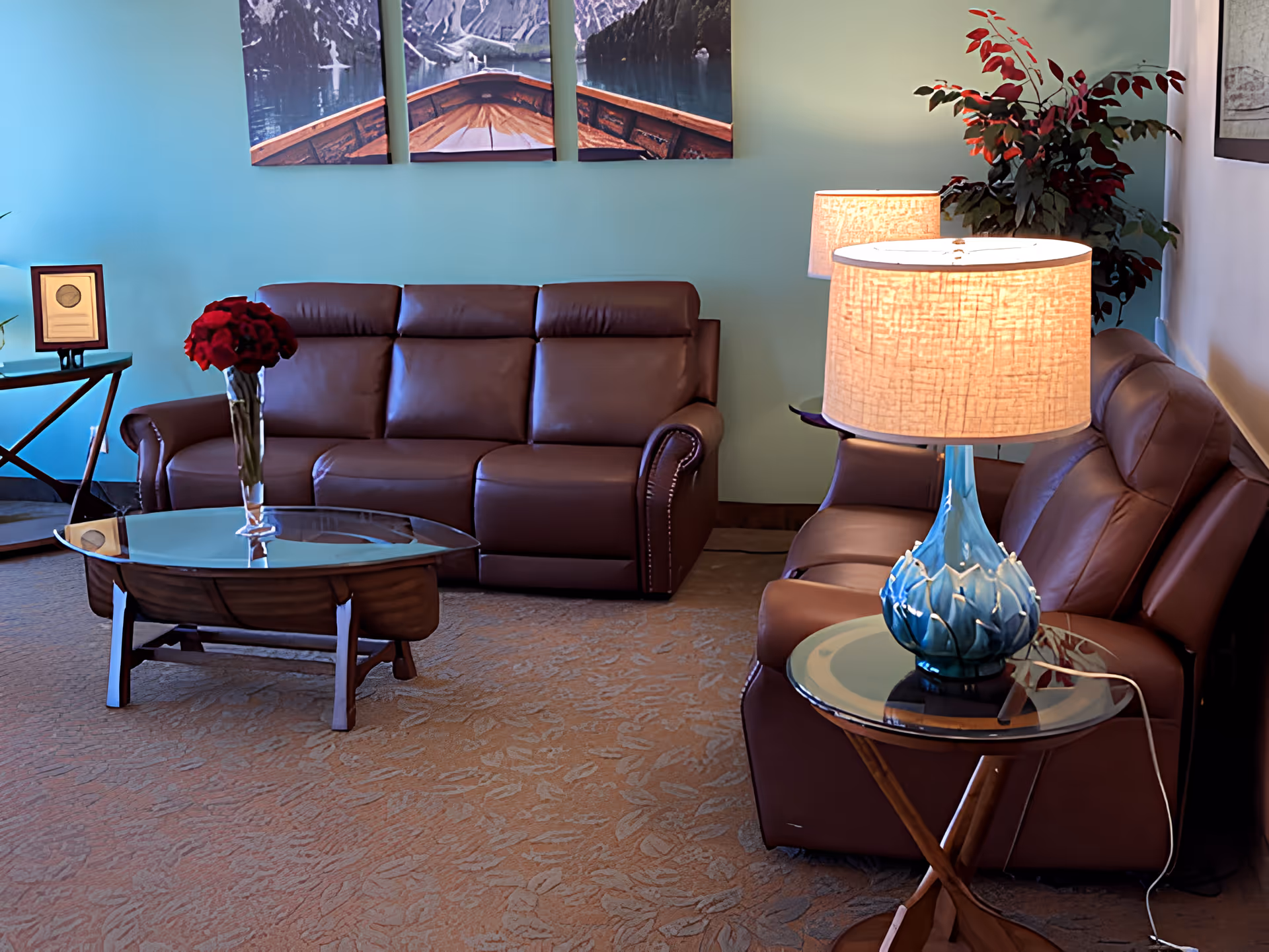 A cozy living room with two brown leather sofas facing each other, a glass coffee table with a vase of red flowers, a side table with a blue decorative lamp, and a green plant in the corner. The walls are painted light blue and decorated with a three-panel artwork depicting a boat on water.