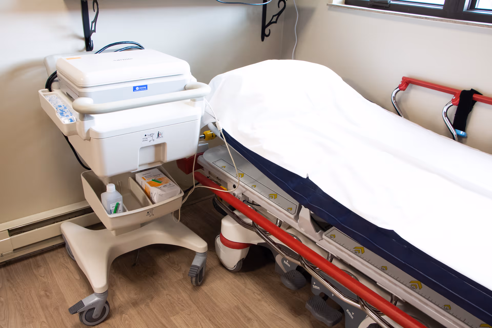 A medical examination room featuring a hospital bed with white sheets and red side rails, next to a white medical cart with various supplies on a wooden floor.