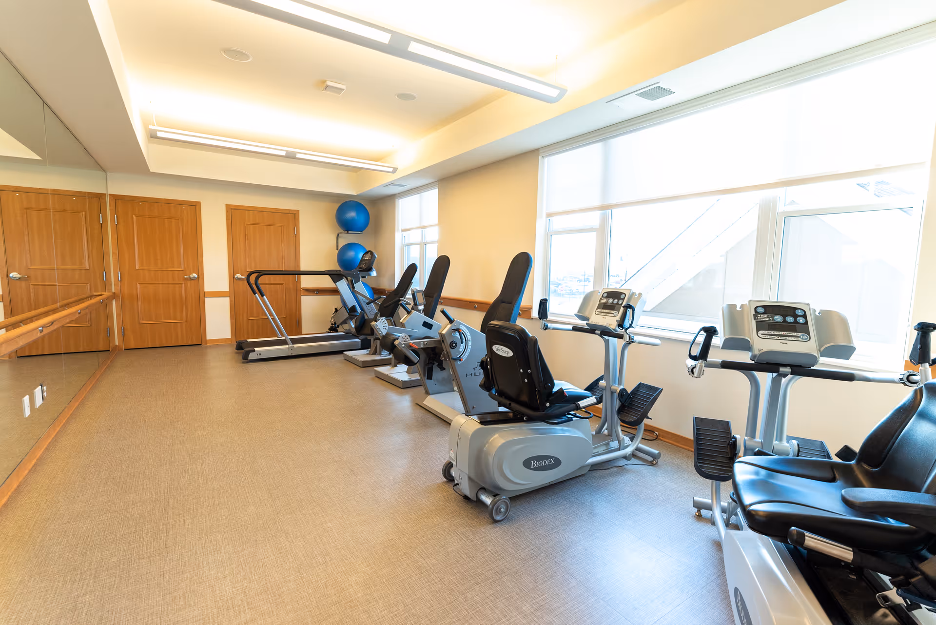 A bright exercise room with several stationary exercise bikes and a treadmill. The room has large windows letting in natural light, wooden doors, a mirrored wall on one side, and two blue exercise balls stacked in the corner.