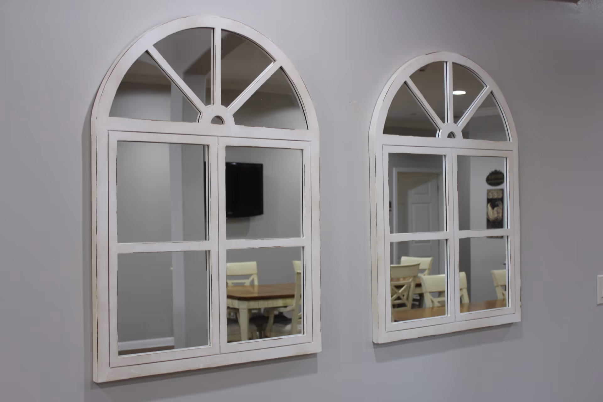 Two white-framed, arched mirrors mounted on a light gray wall reflecting a dining area with a wooden table and cream-colored chairs.