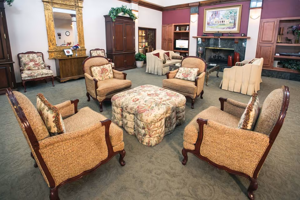 A cozy living room with vintage-style armchairs and floral cushions arranged around a matching floral ottoman. The room features a large ornate gold-framed mirror, a wooden cabinet, a fireplace with a painting above it, and built-in shelves with decorative items. The carpet is green, and the walls have a combination of white and maroon colors.