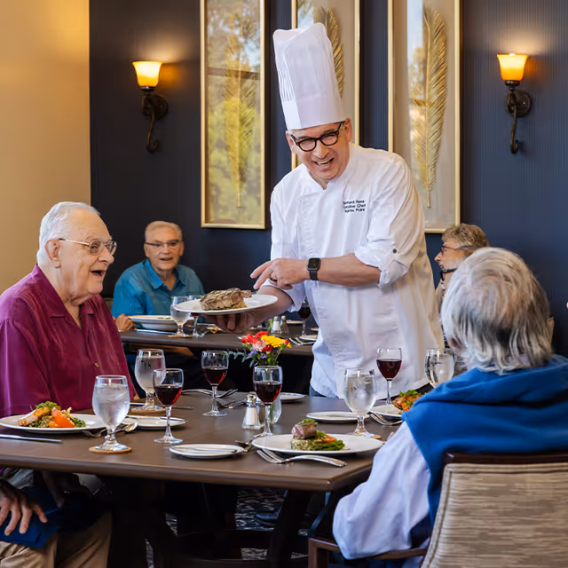 A chef wearing a white uniform and tall chef hat serves a plate of food to a group of elderly people seated around a dining table in a well-lit room with dark walls and decorative wall art. The table is set with plates of food, glasses of water and red wine, and a small flower arrangement.
