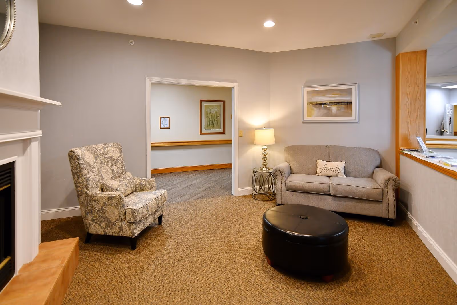 A cozy living room area with a beige sofa that has a pillow labeled 'family', a patterned armchair, a round black ottoman, a side table with a lamp, and a fireplace on the left. The walls are light gray, and there is a framed artwork above the sofa. An open doorway leads to a hallway with framed pictures on the wall.
