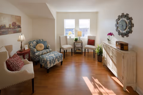 A bright and cozy living room with wooden flooring, featuring two beige armchairs with red and beige cushions, a blue patterned armchair with a matching ottoman, a small red side table with a lamp and picture frame, two white chairs with a small table and lamp between them in front of two windows, a white sideboard with decorative items including a large round wall clock and a potted plant.