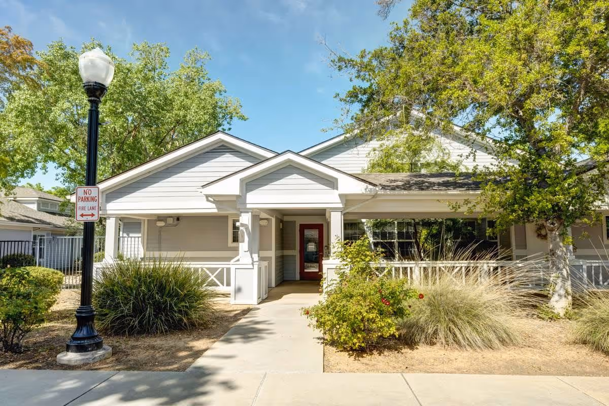 Exterior view of a single-story building with white siding and a covered entrance. There are trees and bushes surrounding the walkway leading to the entrance. A black lamppost with a 'No Parking Fire Lane' sign is visible on the left side. The sky is clear and blue.