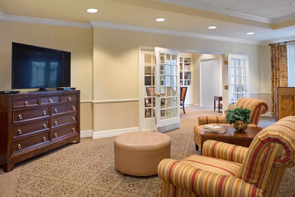A cozy living room area with two striped armchairs, a round beige ottoman, and a wooden coffee table with a potted plant and books. A large flat-screen TV sits on a wooden dresser against a beige wall. French doors open to an adjacent room with bookshelves and chairs. The room has recessed ceiling lights and patterned carpet.
