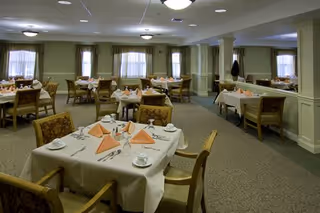 A spacious dining room with multiple tables covered in white tablecloths, each set with folded peach-colored napkins, cups, saucers, and silverware. The room has beige walls, several windows with curtains, and ceiling lights providing illumination. Comfortable upholstered chairs surround the tables.