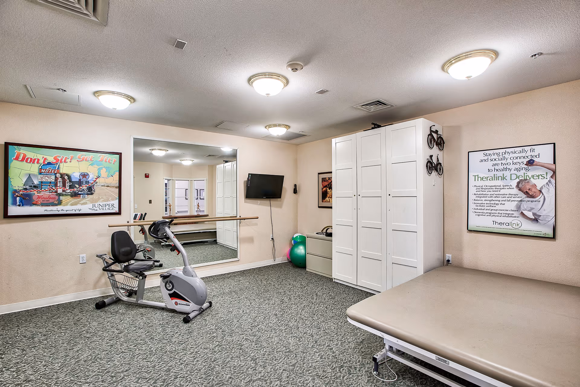 A small exercise room with a recumbent stationary bike, a large wall mirror, a mounted TV, a padded exercise table, and storage cabinets. The room has carpeted flooring and beige walls with two framed posters promoting fitness and healthy aging.