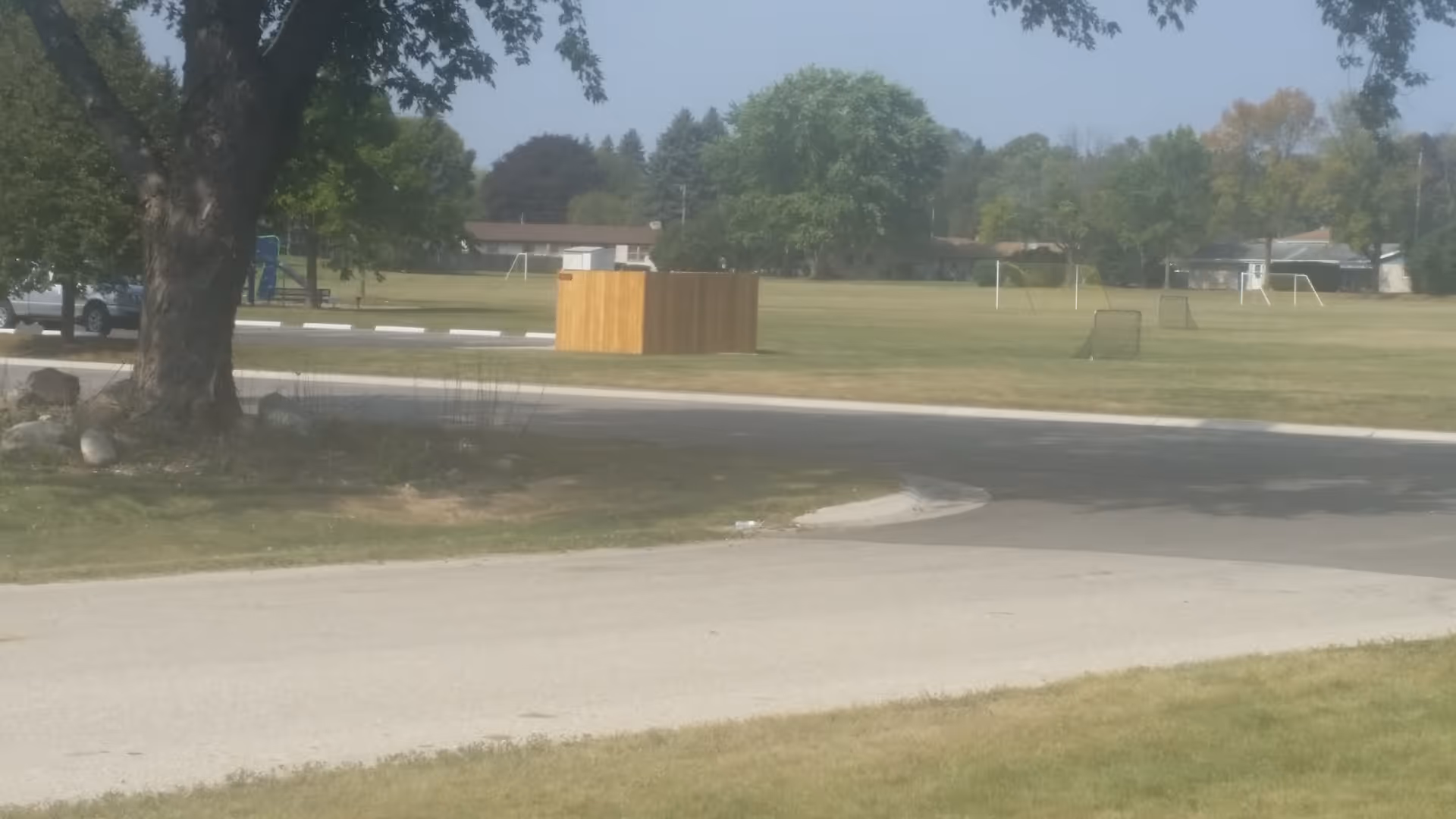 A grassy outdoor area with several soccer goals and a playground in the distance. There are trees and houses in the background, and a paved road runs through the foreground.