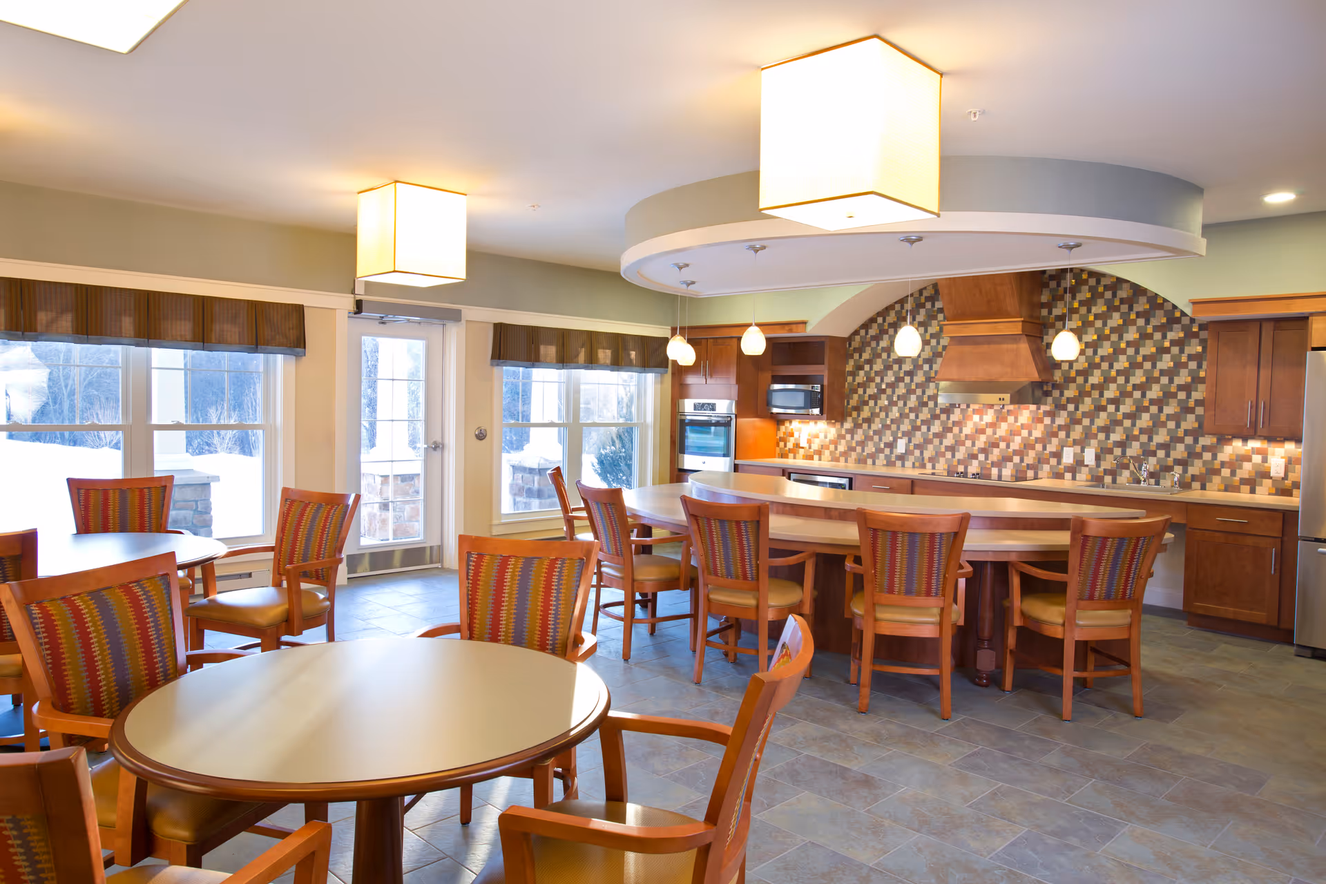 A bright and spacious dining area with round tables and wooden chairs featuring colorful striped cushions. The room includes a large kitchen area with a curved island, wooden cabinets, modern appliances, and a decorative tiled backsplash. Large windows and a glass door let in natural light, and ceiling lights provide additional illumination.